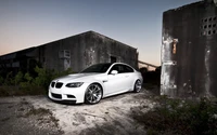 Elegante BMW M3 blanco estacionado junto a edificios desgastados al anochecer, mostrando sus elegantes llantas de aleación y diseño deportivo.