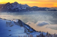 Schneebedeckte Berge unter einem lebhaften Sonnenuntergang mit einem entfernten Bauernhaus, das die ruhige Schönheit einer Winterlandschaft in den Alpen zeigt.