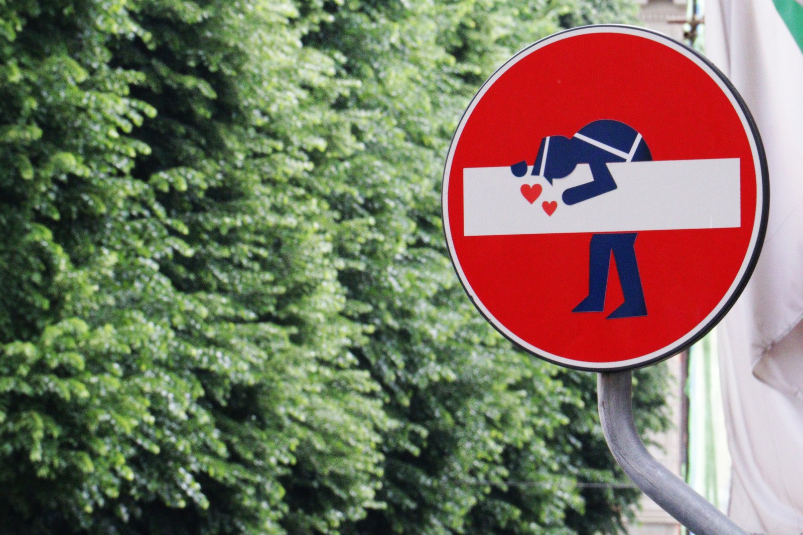 There is a red sign with a man holding a heart on it (signage, green, sign, street sign, tree)