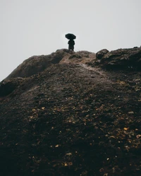 A solitary figure with an umbrella stands atop a misty, rocky hill, surrounded by fog, embodying a sense of solitude and mystery in a dramatic landscape.