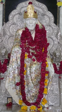 Estátua de Sai Baba adornada com guirlandas florais e uma coroa dourada, sentada em um trono de prata intrincadamente projetado.