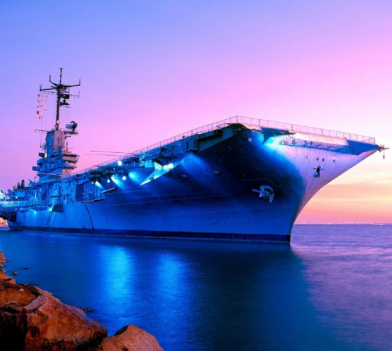 Arafed ship in the water at sunset with rocks in the foreground (aircraft carrier)