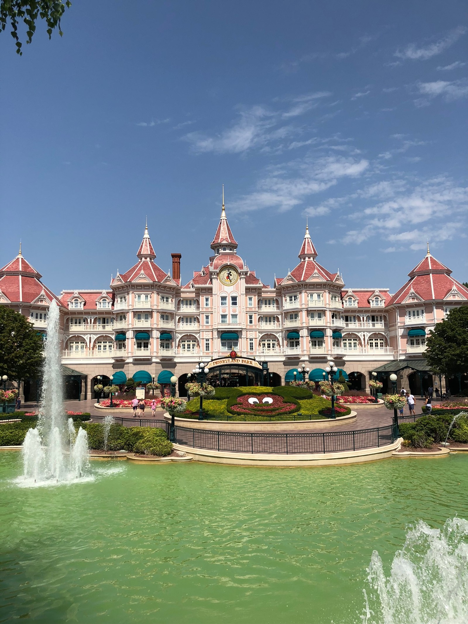 Une fontaine devant un bâtiment avec une tour d'horloge (disneyland, eau, ressources en eau, élément aquatique, point de repère)