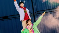 Doyoung y Jungwoo de NCT 127 en atuendos vibrantes, posando de manera juguetona con humo colorido en un fondo urbano.