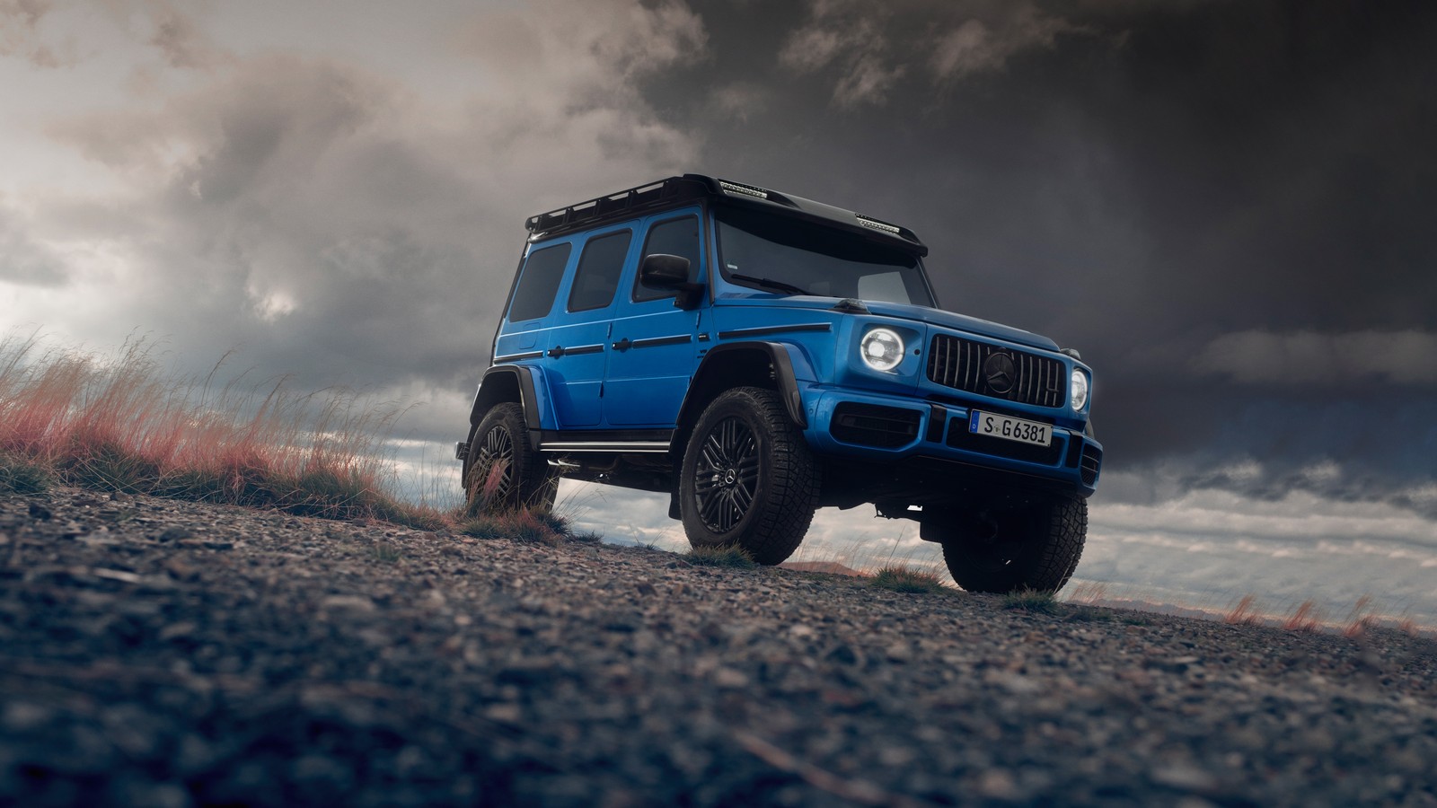 Ein blauer jeep fährt auf einer schotterstraße unter einem bewölkten himmel. (mercedes amg g 63, g wagon, 5k, mercedes amg g klasse suv, mercedes amg g class suv)