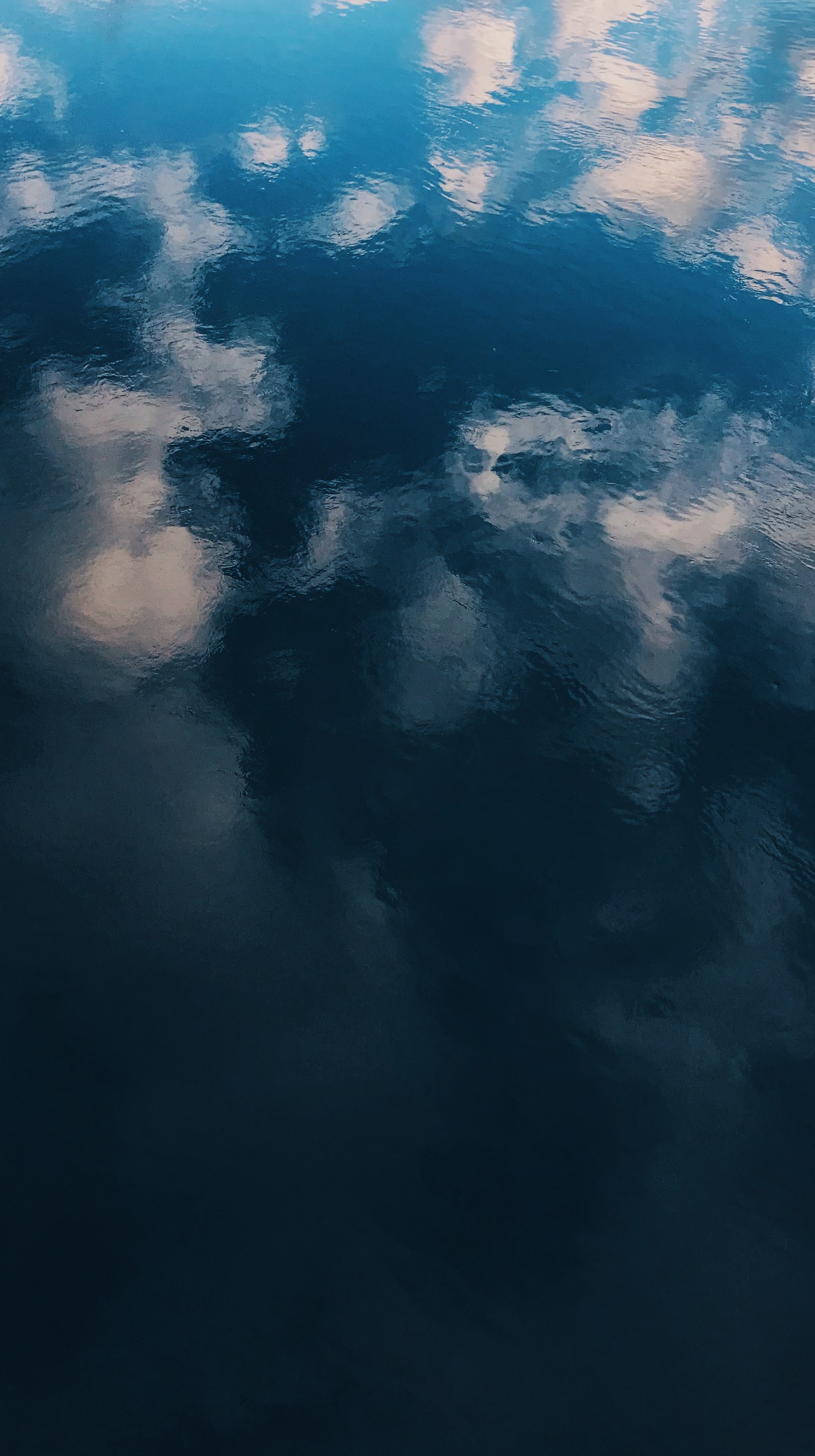 Um barco flutuando na água com nuvens refletidas nele (preto, azul, nuvens, água, reflexo)