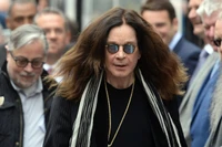 Ozzy Osbourne com cabelo longo e óculos de sol, vestido com um traje preto estiloso, exalando uma vibração clássica de estrela do rock em meio a uma multidão.