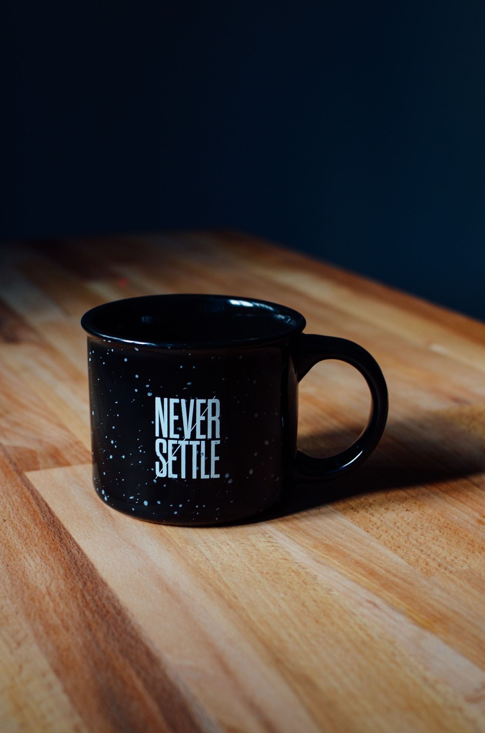 There is a black coffee mug sitting on a wooden table (quotes, never settle, motivational, mood, tech)