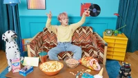 Yeonjun von Tomorrow X Together genießt einen nostalgischen Moment in einem bunten, retro-inspirierten Wohnzimmer, das mit Snacks und verspielter Dekoration gefüllt ist.