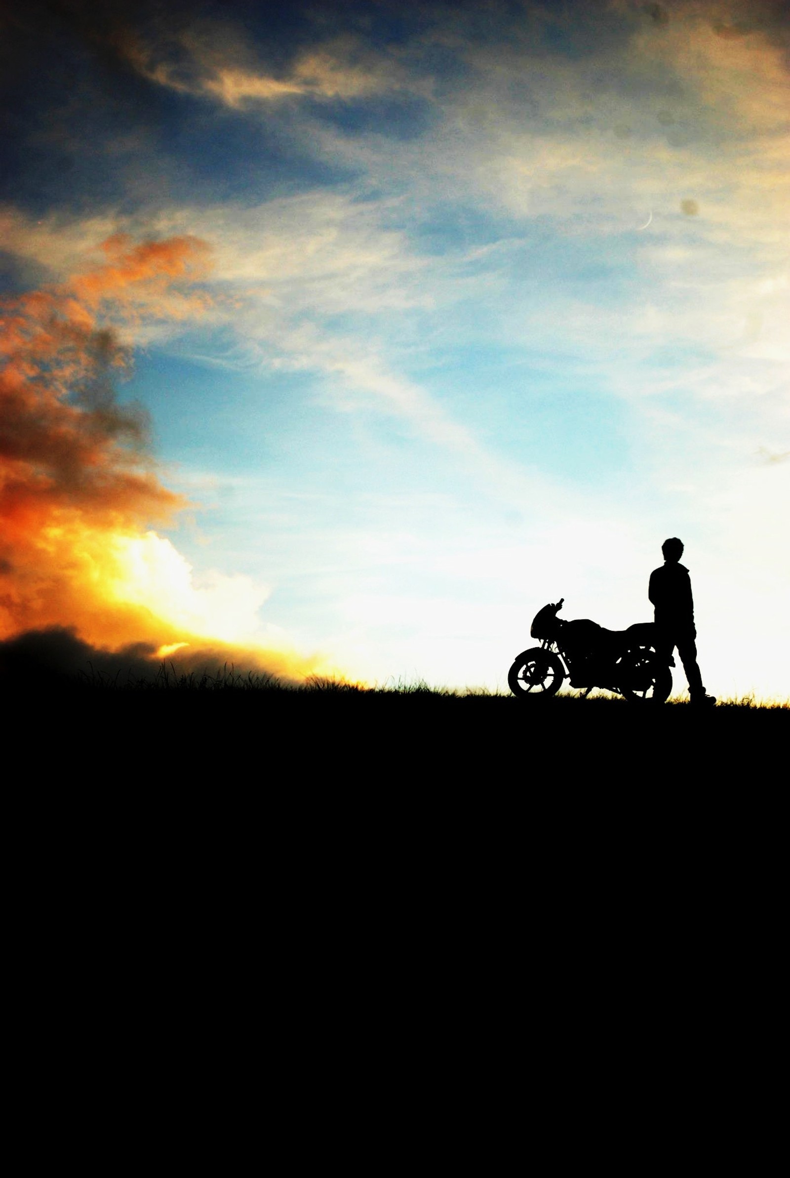 There is a man standing next to a motorcycle on a hill (bike, clouds)