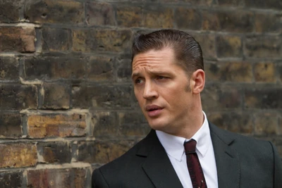 Tom Hardy en tant qu'homme d'affaires sophistiqué dans un costume sur mesure, dégageant du charisme sur un fond de briques texturées.