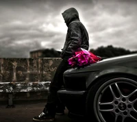 Un homme en hoodie attend à côté d'une voiture, tenant un bouquet de fleurs vibrantes.