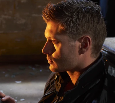 Dean Winchester dans une pièce faiblement éclairée, montrant son regard intense et son charme rugueux.