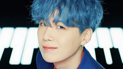 Suga of BTS with vibrant blue hair, smiling against a dynamic background.