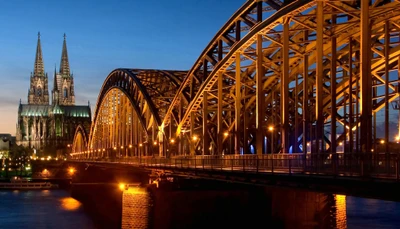 Puente iluminado por la noche con reflejos, con la Catedral de Colonia al fondo, mostrando un impresionante paisaje urbano sobre el Rin.