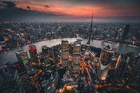 Une vue aérienne époustouflante de la skyline illuminée de Shanghai au crépuscule, mettant en valeur les gratte-ciels de la ville le long de la rivière, incarnant l'élégance urbaine et la vitalité métropolitaine.