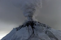 Estratovulcão em erupção com nuvens de cinzas contra um céu nublado, mostrando atividade geológica e terreno montanhoso.