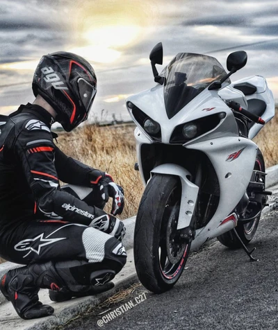 Un motard en tenue admire une moto sportive blanche élégante contre un ciel dramatique au coucher du soleil.