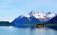 Montanhas cobertas de neve se erguem majestosas acima de um fiorde sereno, emolduradas por um céu azul claro, exibindo a beleza pristina da natureza selvagem do Alasca.