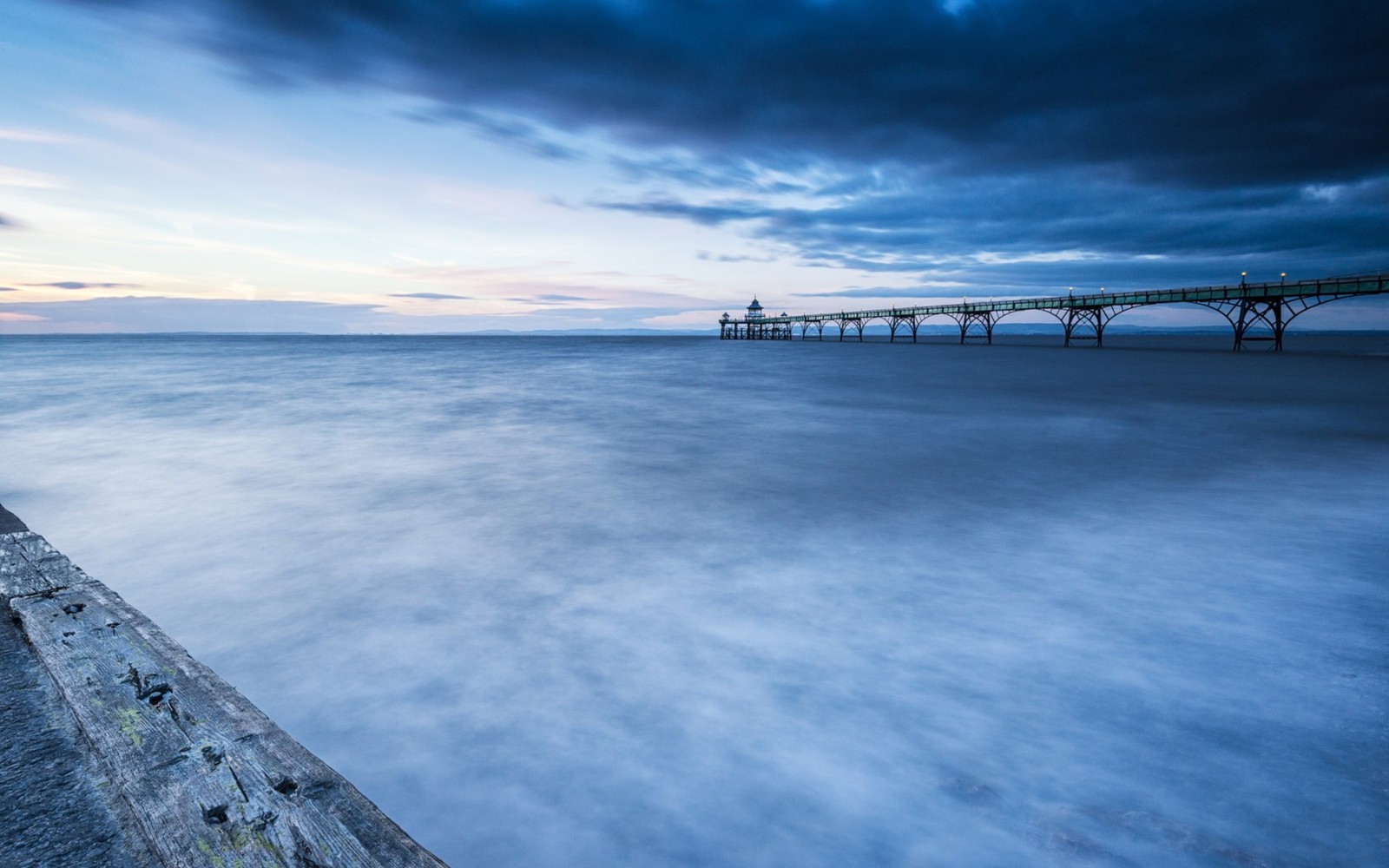 A long exposure photograph of a pier at dusk with a cloudy sky (blue, bridge, calm, clouds, cool)