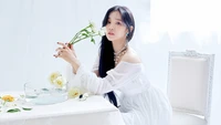 Song Yuqi dans une robe blanche éthérée, tenant une rose, entourée de fleurs délicates et d'une lumière douce, incarnant l'élégance et la sérénité.