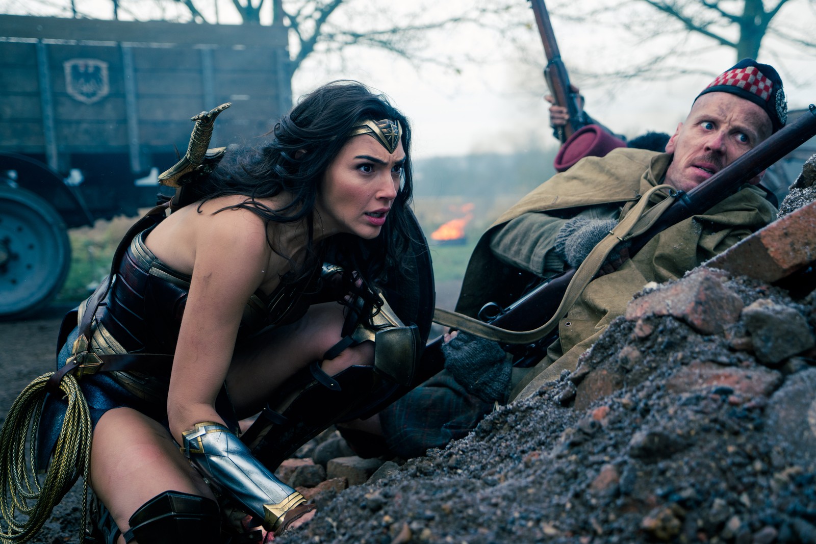 Une femme arabe en costume à genoux à côté d'un homme en trench (wonder woman, gal gadot, arbre, dc comics, film de super héros)