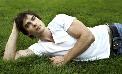 Ian Somerhalder en tant que Damon Salvatore, allongé sur une pelouse verdoyante dans un t-shirt blanc décontracté, dégageant une ambiance estivale.