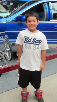 Un niño sonriente con una camiseta blanca de Old Navy está de pie frente a un coche lowrider azul en una convención de 2015 en Phoenix.