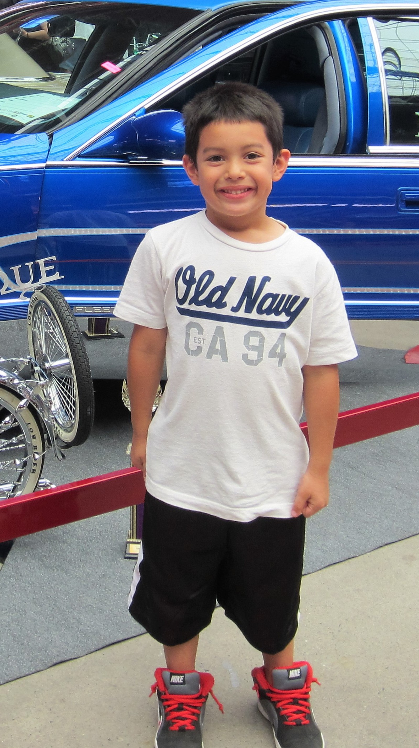 Un niño árabe con una camisa blanca y pantalones cortos negros de pie frente a un camión azul (lowerider show 2015, phoenix convention)