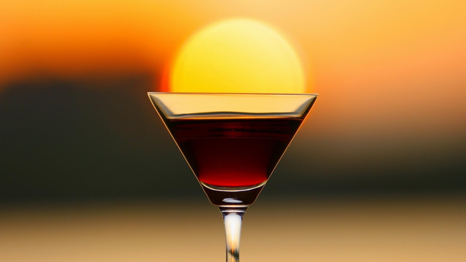 There is a glass of wine sitting on a table with a sunset in the background (drink, classic cocktail, alcoholic beverage, distilled beverage, manhattan)