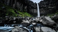 Cascata majestosa caindo sobre penhascos acidentados, cercada por vegetação exuberante e terreno rochoso.