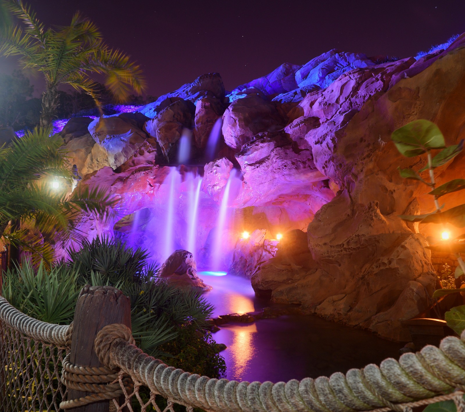 There is a waterfall that is lit up at night with colorful lights (landscape, nature, oasis, palm trees, tropical)