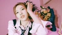 Sana from TWICE poses elegantly against a vibrant pink backdrop, showcasing a stylish outfit and captivating expression, with decorative elements like flowers and a statue enhancing the aesthetic.