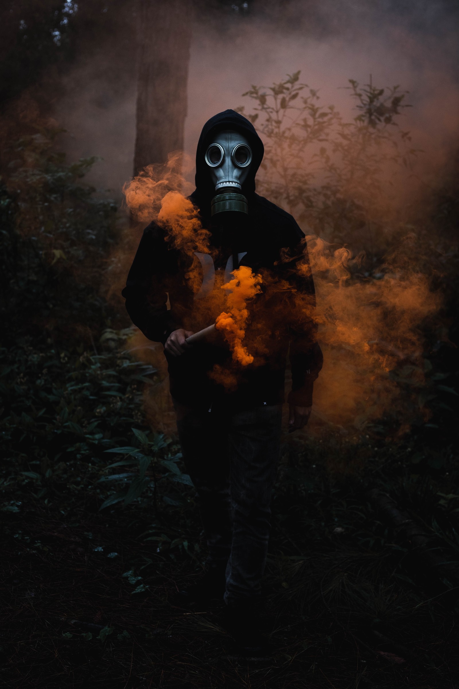 Homme retravaillé en masque à gaz tenant un bâton dans une forêt (homme masqué, veste noire, smoke can, arrière plan sombre, fumée orange)