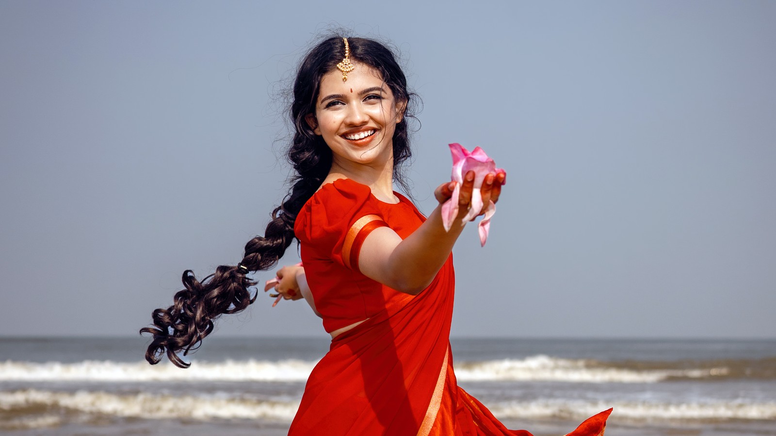 Une femme en robe rouge tenant une fleur rose sur la plage. (mamitha baiju, 5k, actrice malayalam, actrice indienne, sari)