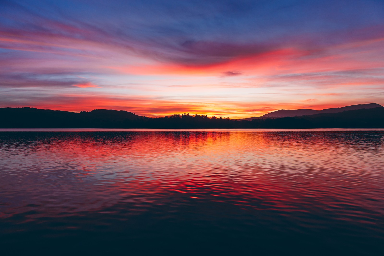 Um pôr do sol vermelho e azul sobre um lago com montanhas ao fundo (por do sol, horizonte, nascer do sol, corpo de água, resplendor)