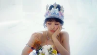 Wendy from Red Velvet in a dreamy setting, wearing a crown, lost in thought with a whimsical bouquet.