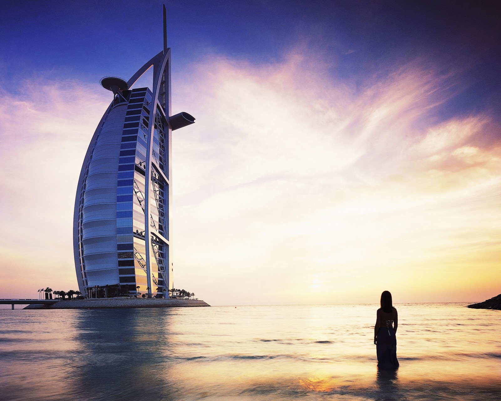 Uma girafa em pé na água em frente a um prédio alto (burj al arab, por do sol, horizonte, mar, suíte)