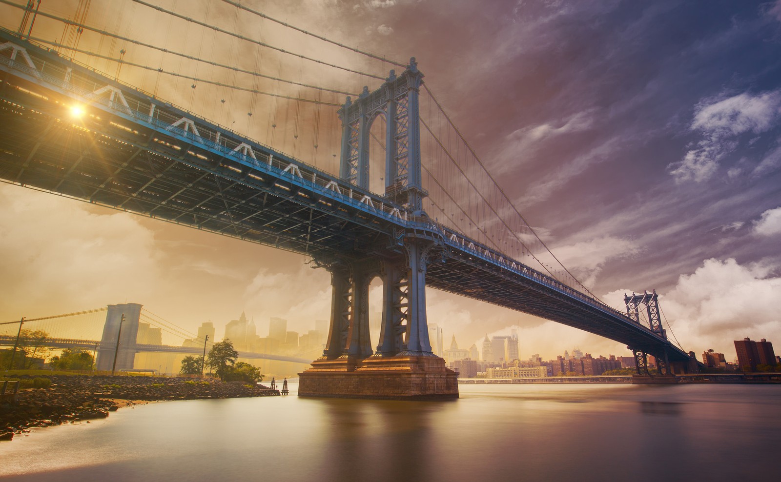 Uma vista de uma ponte sobre um corpo d'água com uma cidade ao fundo. (ponte de manhattan, manhattan bridge, ponte suspensa, ponte, marco)