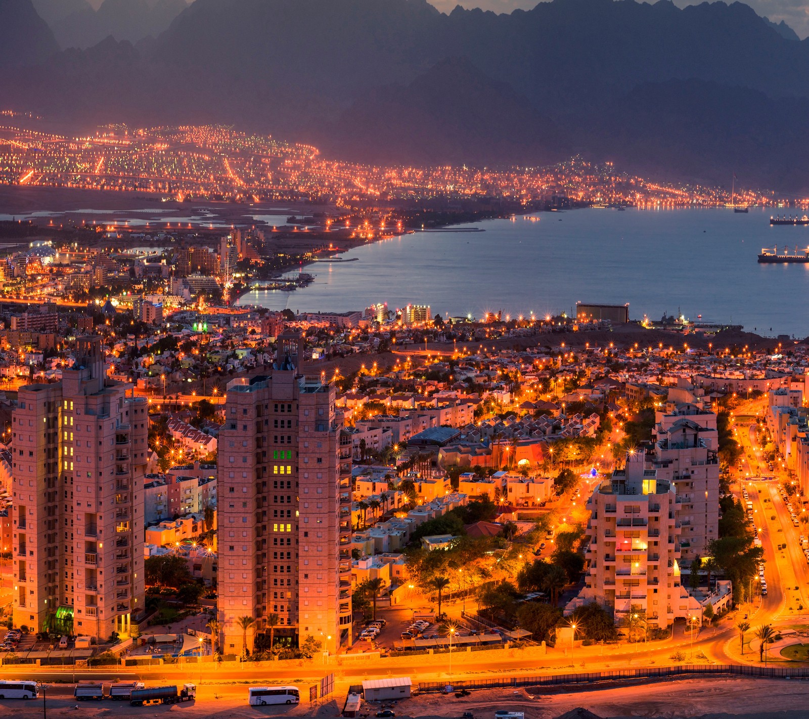 Uma vista aérea de uma cidade à noite com um navio de cruzeiro ao longe (cidade, israel, israelense, noite, urbano)