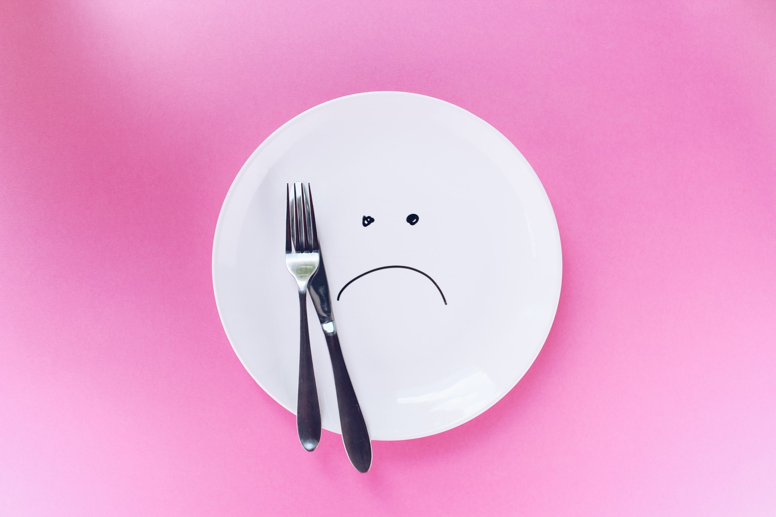 Araffe plate with a sad face drawn on it with a fork and knife (empty plate, sad, fork, pink background, hunger)