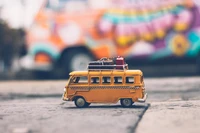 Yellow Volkswagen Type 2 Van Model with Luggage on Rooftop Against a Colorful Background.