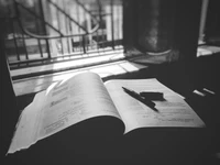 Open book on a table near a window, with a pen resting beside it, illuminated by soft sunlight.