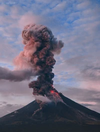 Vulcão escudo em erupção liberando fumaça escura e lava na atmosfera contra um céu nublado.