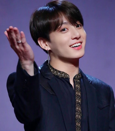 Jungkook do BTS, sorrindo calorosamente e gesticulando com a mão enquanto usa um traje preto estiloso.