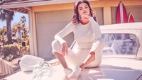 Selena Gomez dans une tenue blanche élégante, posant sur une voiture vintage devant un arrière-plan maritime vibrant.