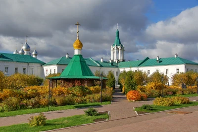 Historic monastery with a green spire and golden cross, surrounded by vibrant shrubland and trees under a cloudy sky.