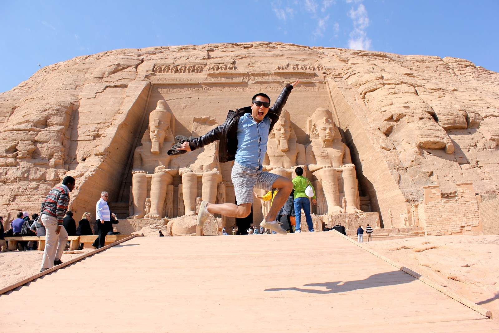 Um homem pulando em frente a um grande edifício de pedra (assuã, viagem, luxor, turismo, sítio histórico)