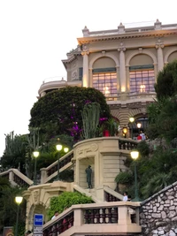 Fachada del Casino de Montecarlo con exuberante vegetación, escaleras elegantes y iluminación decorativa.