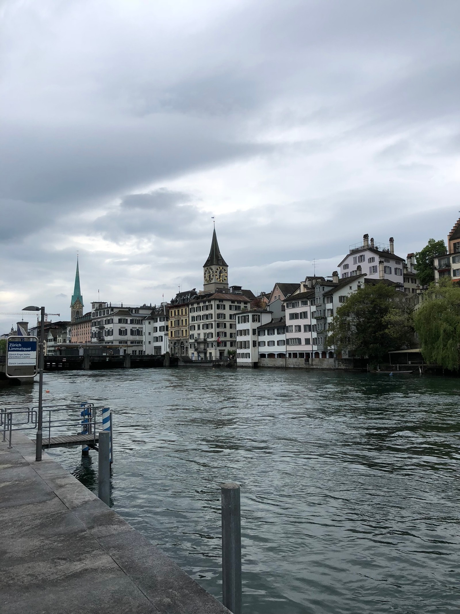 There is a boat that is going down the river in the city (limmat, water, waterway, cloud, town)